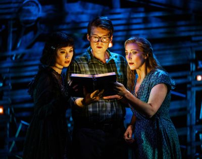 Three people stand under dramatic stage lighting, intensely reading a book. The scene conveys mystery and intrigue with a dark, theatrical backdrop.