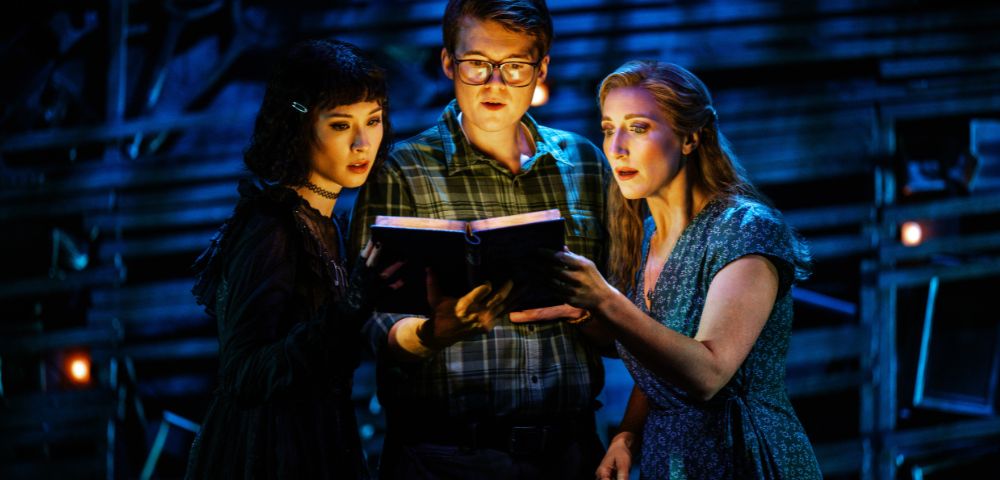 Three people stand under dramatic stage lighting, intensely reading a book. The scene conveys mystery and intrigue with a dark, theatrical backdrop.