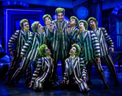 A group of performers in green wigs and striped suits pose energetically on stage, creating a theatrical and lively atmosphere with dramatic expressions.