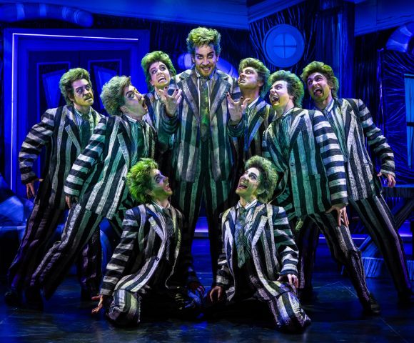 A group of performers in green wigs and striped suits pose energetically on stage, creating a theatrical and lively atmosphere with dramatic expressions.