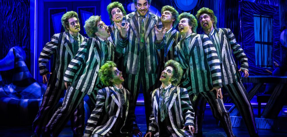 A group of performers in green wigs and striped suits pose energetically on stage, creating a theatrical and lively atmosphere with dramatic expressions.
