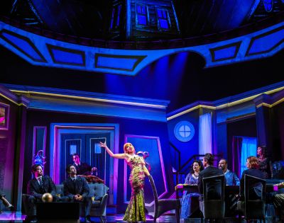 A lively stage scene with a woman in a sparkling dress singing, surrounded by seated figures in a vibrant, dramatic setting with vivid blue and purple lighting.