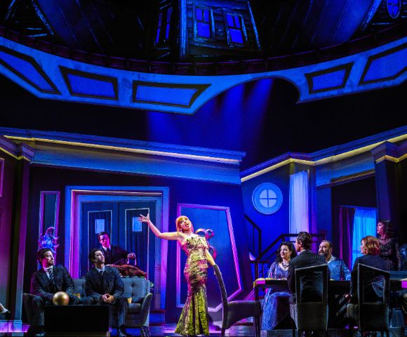 A lively stage scene with a woman in a sparkling dress singing, surrounded by seated figures in a vibrant, dramatic setting with vivid blue and purple lighting.