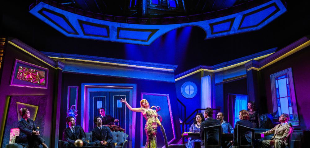 A lively stage scene with a woman in a sparkling dress singing, surrounded by seated figures in a vibrant, dramatic setting with vivid blue and purple lighting.