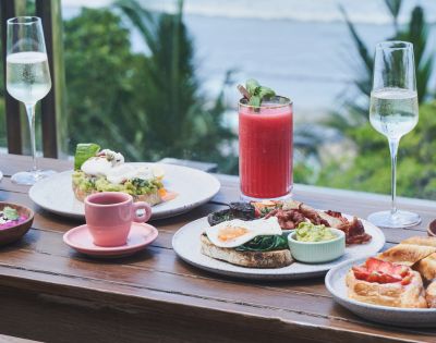 Outdoor breakfast spread with avocado toast, poached eggs, and bacon. Includes a pink smoothie, coffee, and sparkling drinks. Ocean view backdrop.