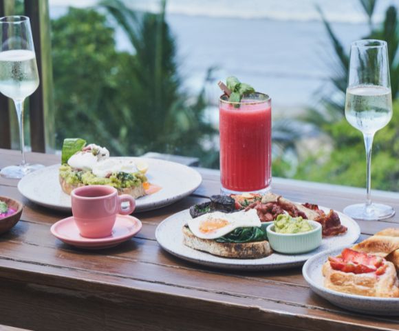 Outdoor breakfast spread with avocado toast, poached eggs, and bacon. Includes a pink smoothie, coffee, and sparkling drinks. Ocean view backdrop.