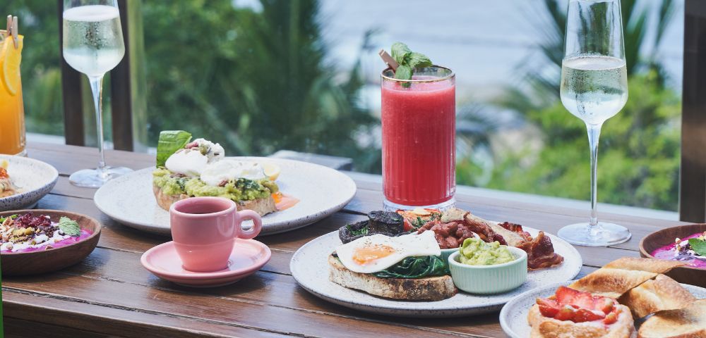 Outdoor breakfast spread with avocado toast, poached eggs, and bacon. Includes a pink smoothie, coffee, and sparkling drinks. Ocean view backdrop.