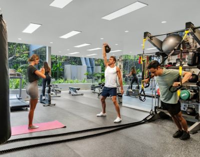Three people exercise in a modern gym. One lifts a kettlebell, another uses TRX bands, and a woman stands on a yoga mat. Bright, open space, focused atmosphere.