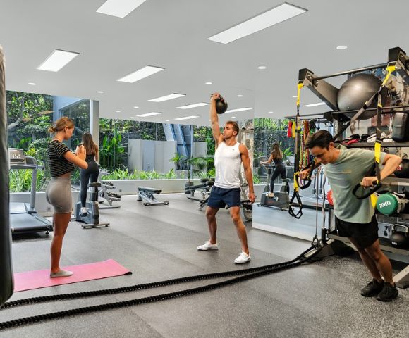 Three people exercise in a modern gym. One lifts a kettlebell, another uses TRX bands, and a woman stands on a yoga mat. Bright, open space, focused atmosphere.