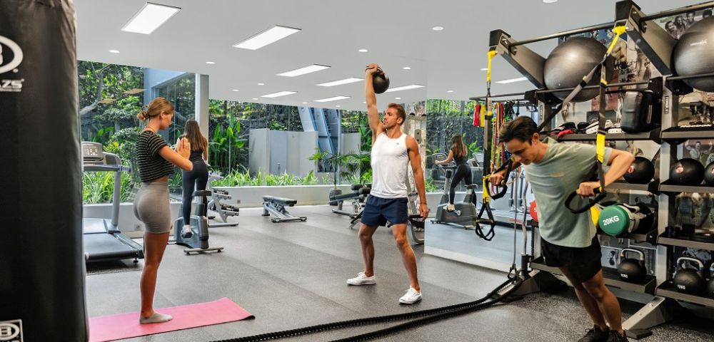 Three people exercise in a modern gym. One lifts a kettlebell, another uses TRX bands, and a woman stands on a yoga mat. Bright, open space, focused atmosphere.
