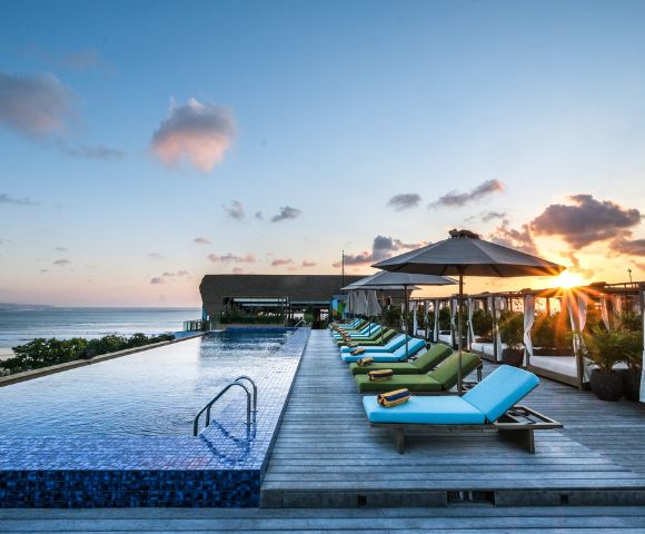 Rooftop infinity pool at sunset with vibrant lounge chairs and umbrellas, ocean view in the background, creating a serene and luxurious atmosphere.