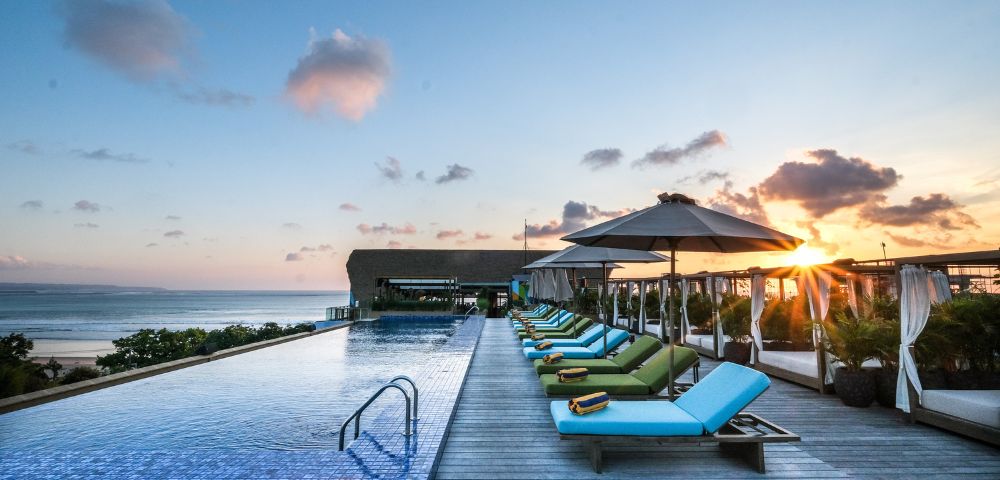 Rooftop infinity pool at sunset with vibrant lounge chairs and umbrellas, ocean view in the background, creating a serene and luxurious atmosphere.