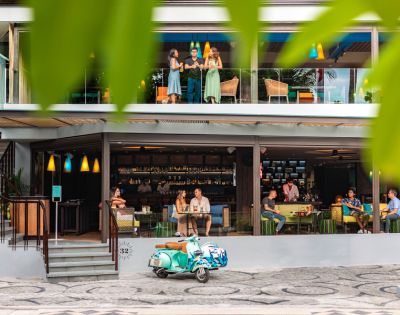A vibrant café with a colorful Vespa parked outside. Patrons enjoy drinks inside and on the balcony. Green leaves frame the lively scene.