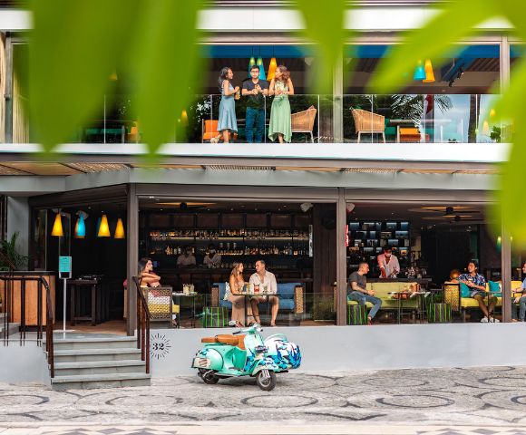 A vibrant café with a colorful Vespa parked outside. Patrons enjoy drinks inside and on the balcony. Green leaves frame the lively scene.