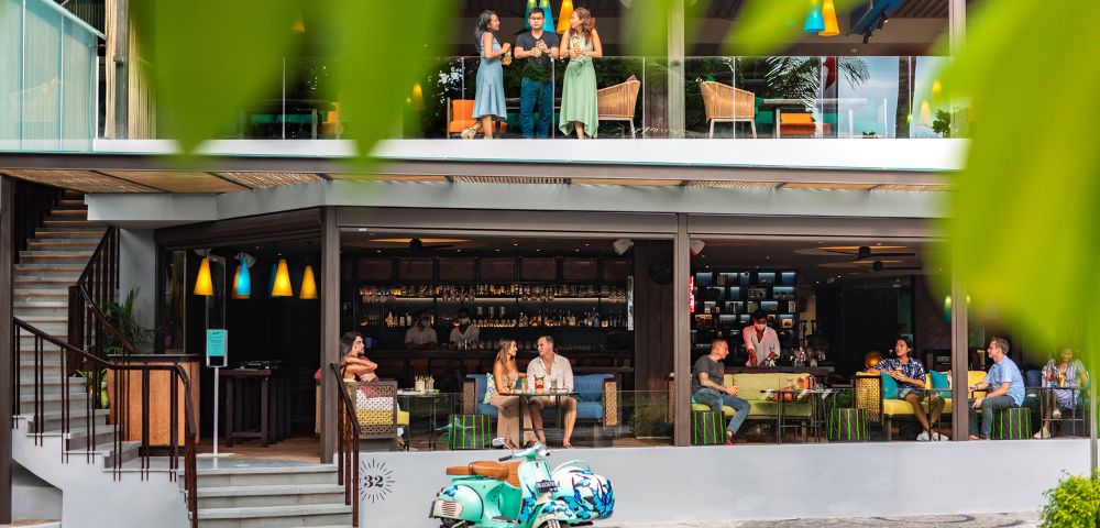 A vibrant café with a colorful Vespa parked outside. Patrons enjoy drinks inside and on the balcony. Green leaves frame the lively scene.