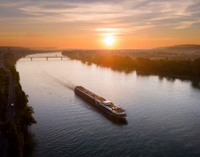 A long cruise ship sails down a wide river under a vibrant sunset sky. The warm glow reflects off the water, conveying tranquility and serenity.