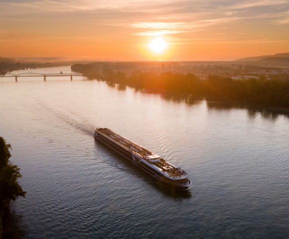 A long cruise ship sails down a wide river under a vibrant sunset sky. The warm glow reflects off the water, conveying tranquility and serenity.