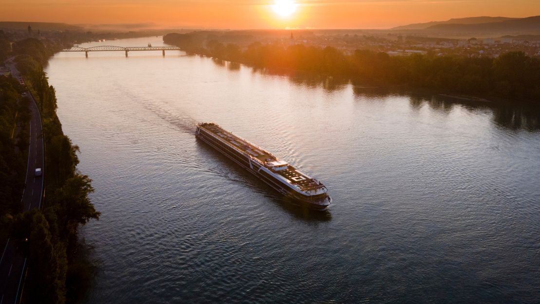A long cruise ship sails down a wide river under a vibrant sunset sky. The warm glow reflects off the water, conveying tranquility and serenity.