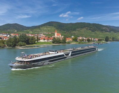 A sleek river cruise ship sails along a calm river, with a picturesque village and green hills under a clear blue sky in the background.