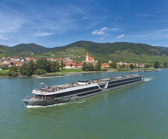 A sleek river cruise ship sails along a calm river, with a picturesque village and green hills under a clear blue sky in the background.