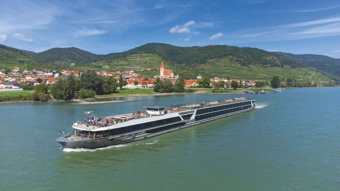 A sleek river cruise ship sails along a calm river, with a picturesque village and green hills under a clear blue sky in the background.