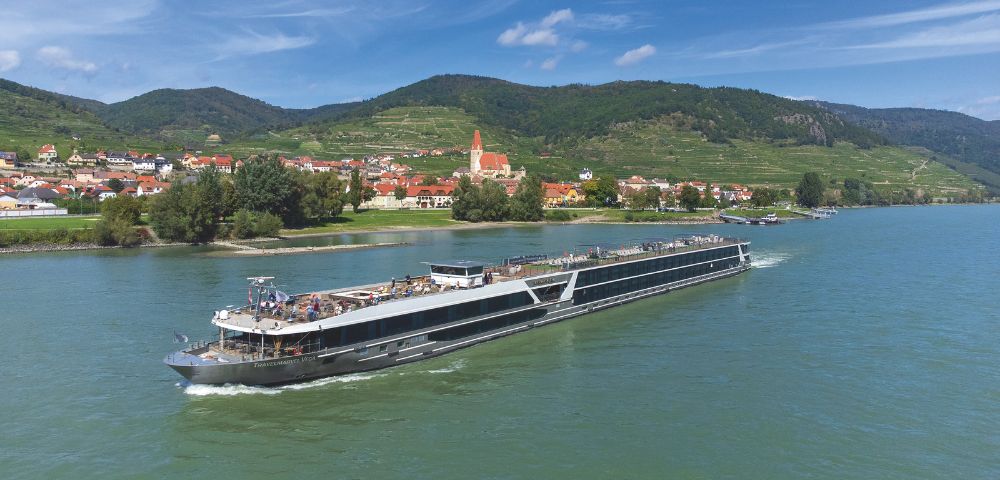 A sleek river cruise ship sails along a calm river, with a picturesque village and green hills under a clear blue sky in the background.