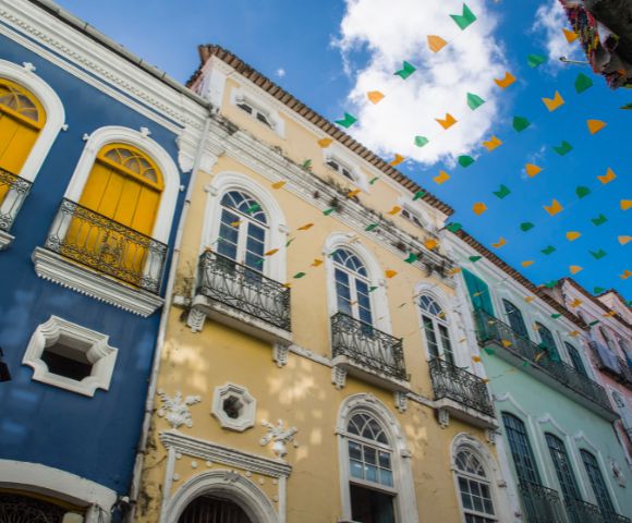 Colorful colonial buildings with arched windows and intricate iron balconies are adorned with strings of festive green and orange pennants under a blue sky.