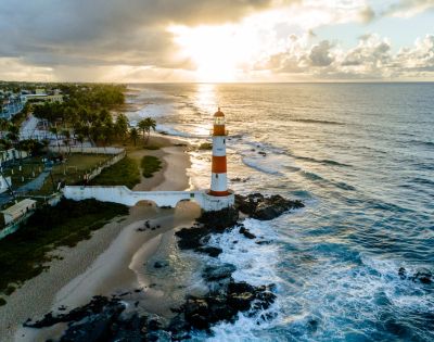 Aerial view of a striped lighthouse on a rocky shore at sunrise, surrounded by ocean waves and a sandy beach. Palm trees and houses line the distant coast.