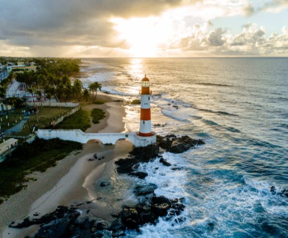 Aerial view of a striped lighthouse on a rocky shore at sunrise, surrounded by ocean waves and a sandy beach. Palm trees and houses line the distant coast.
