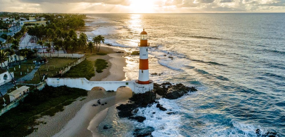 Aerial view of a striped lighthouse on a rocky shore at sunrise, surrounded by ocean waves and a sandy beach. Palm trees and houses line the distant coast.