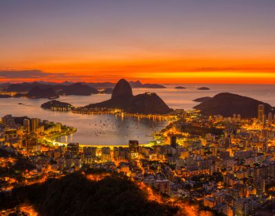 A stunning aerial view of Rio de Janeiro at sunset, with vibrant orange skies illuminating Sugarloaf Mountain and the cityscape below, creating a warm, serene atmosphere.