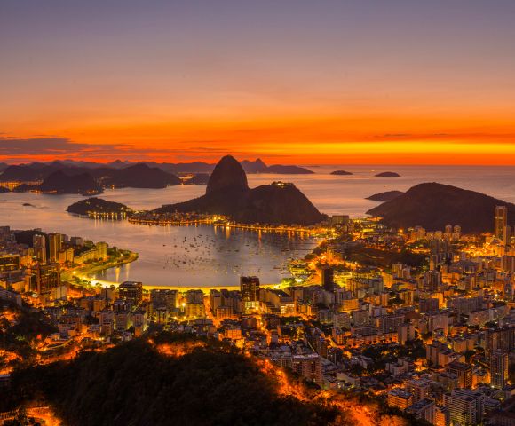 A stunning aerial view of Rio de Janeiro at sunset, with vibrant orange skies illuminating Sugarloaf Mountain and the cityscape below, creating a warm, serene atmosphere.