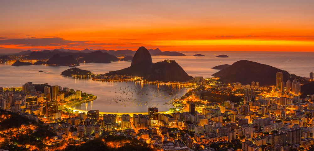 A stunning aerial view of Rio de Janeiro at sunset, with vibrant orange skies illuminating Sugarloaf Mountain and the cityscape below, creating a warm, serene atmosphere.