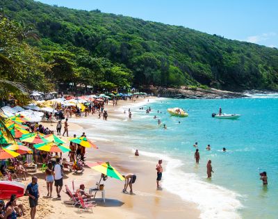 A lively beach scene with colorful umbrellas, people sunbathing and swimming. Lush green hills in the background, clear blue sky overhead. Energetic and vibrant.
