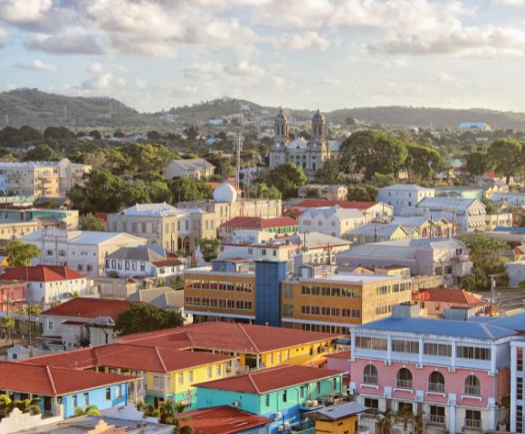 Aerial view of a colorful town with pastel buildings, red roofs, and lush green trees under a partly cloudy sky, conveying a peaceful, vibrant atmosphere.