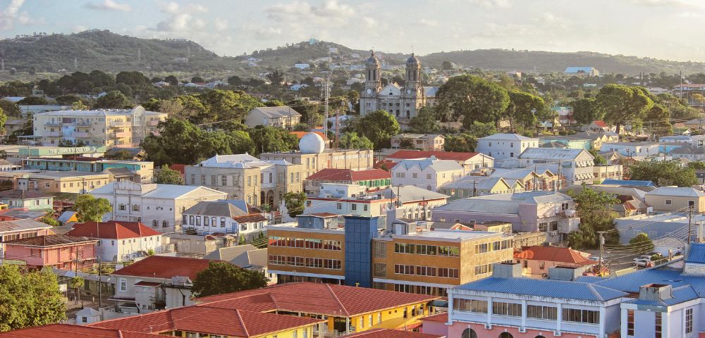 Aerial view of a colorful town with pastel buildings, red roofs, and lush green trees under a partly cloudy sky, conveying a peaceful, vibrant atmosphere.