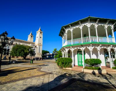 A sunlit plaza features a white gazebo with green accents and a historic church with twin towers. Blue sky and trees complete the serene scene.