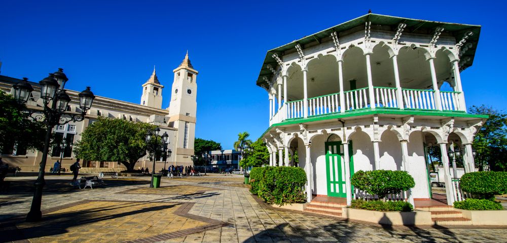 A sunlit plaza features a white gazebo with green accents and a historic church with twin towers. Blue sky and trees complete the serene scene.