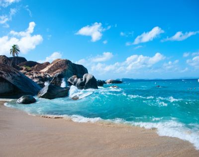 Tropical beach with clear turquoise water, gentle waves, tan sand, and large rocks. A palm tree stands on rocks under a bright blue sky with fluffy clouds.