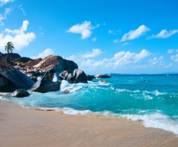 Tropical beach with clear turquoise water, gentle waves, tan sand, and large rocks. A palm tree stands on rocks under a bright blue sky with fluffy clouds.