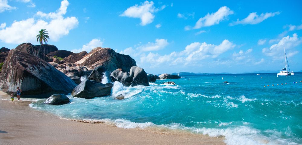 Tropical beach with clear turquoise water, gentle waves, tan sand, and large rocks. A palm tree stands on rocks under a bright blue sky with fluffy clouds.