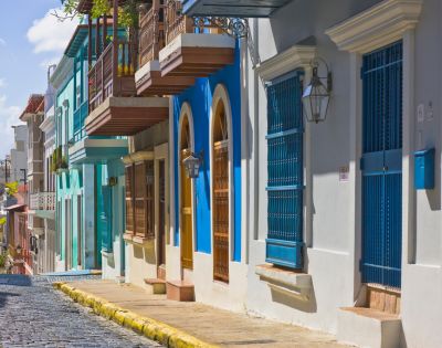 Colorful, historic street with vibrant blue, green, and white buildings featuring wooden balconies and iron grilles. Sunny day on a cobblestone road.