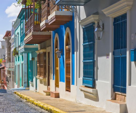 Colorful, historic street with vibrant blue, green, and white buildings featuring wooden balconies and iron grilles. Sunny day on a cobblestone road.