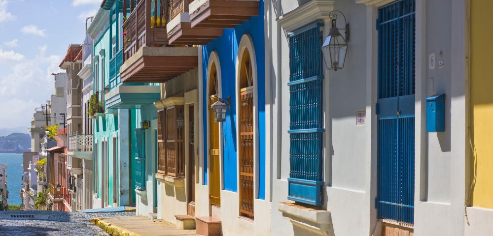 Colorful, historic street with vibrant blue, green, and white buildings featuring wooden balconies and iron grilles. Sunny day on a cobblestone road.