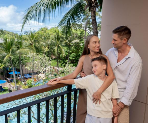 A family of three stands on a balcony overlooking a lush tropical garden and pool. The parents smile at each other while holding their son, conveying warmth and contentment.