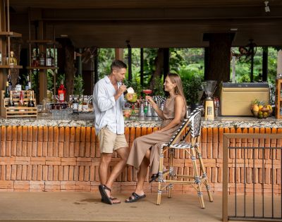 A couple enjoys drinks at a stylish outdoor bar with a brick counter and lush greenery in the background, creating a relaxed, tropical vibe.