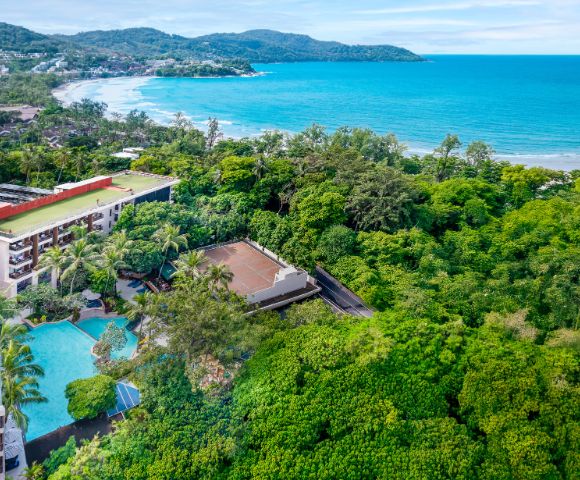 Aerial view of a coastal resort surrounded by lush greenery, with a swimming pool and tennis court in the foreground, and a turquoise ocean and sandy beach in the background, conveying a serene and tropical getaway.