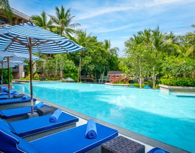 Tropical pool scene with blue lounge chairs and striped umbrellas along a calm pool. Surrounded by lush green palm trees, under a clear blue sky.