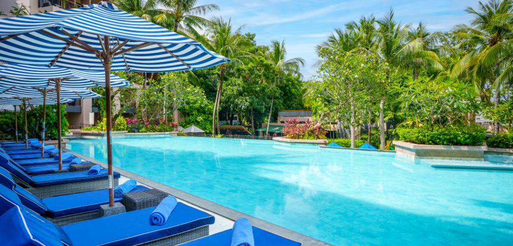 Tropical pool scene with blue lounge chairs and striped umbrellas along a calm pool. Surrounded by lush green palm trees, under a clear blue sky.