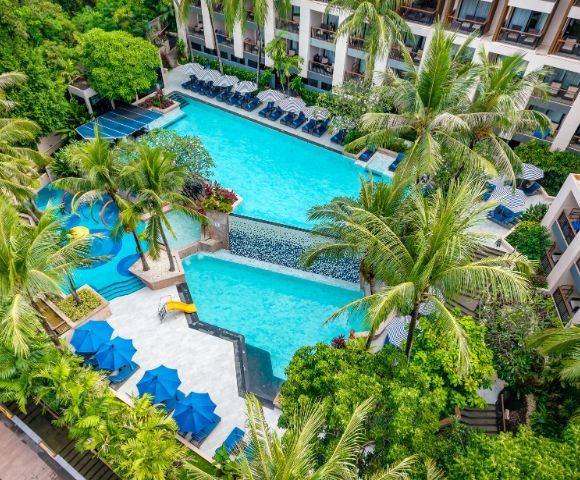 Aerial view of a resort's vibrant pool area, surrounded by lush greenery and palm trees. Blue sun umbrellas and lounge chairs add a relaxing, tropical vibe.